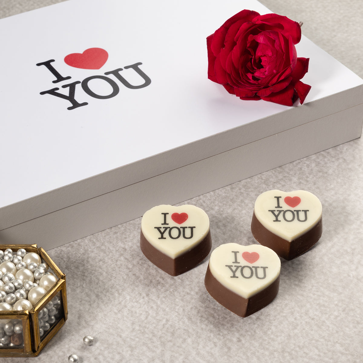 Heart-shaped chocolates with 'I ❤️ YOU' text, a red rose, and 'I ❤️ YOU' card on a textured surface.