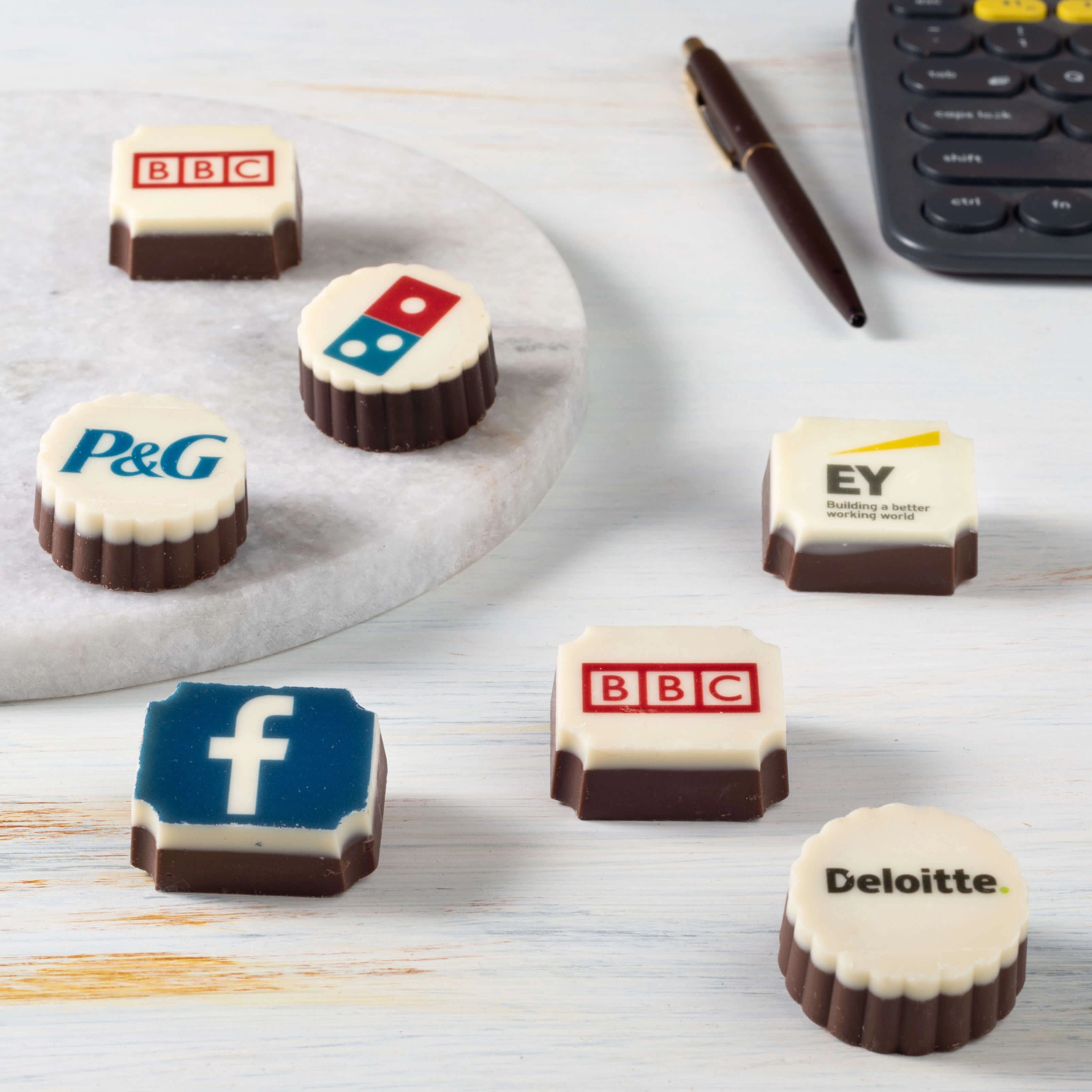 Close‑up logo chocolates for Facebook, EY, P&G, Domino’s on marble desktop—ideal upscale office gifting snack.